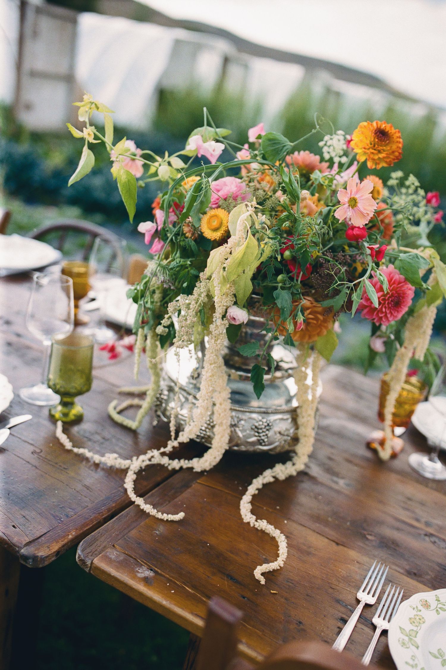 Rustic wooden table with a vibrant wildflower centerpiece in a silver vase, draped with pearl garlands at an outdoor wedding.