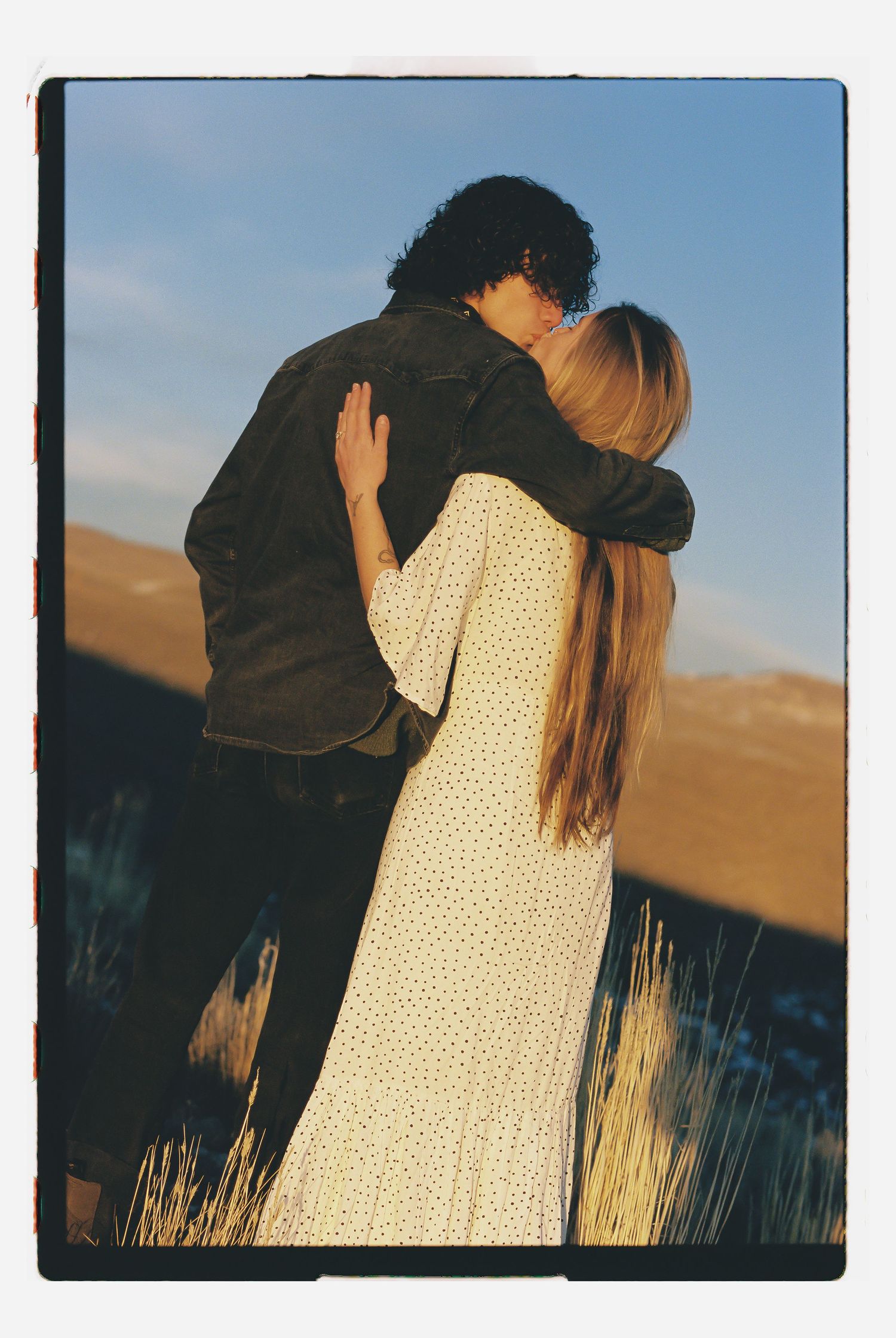 Couple sharing a romantic kiss outdoors at golden hour, woman in floral dress embracing man in denim jacket.