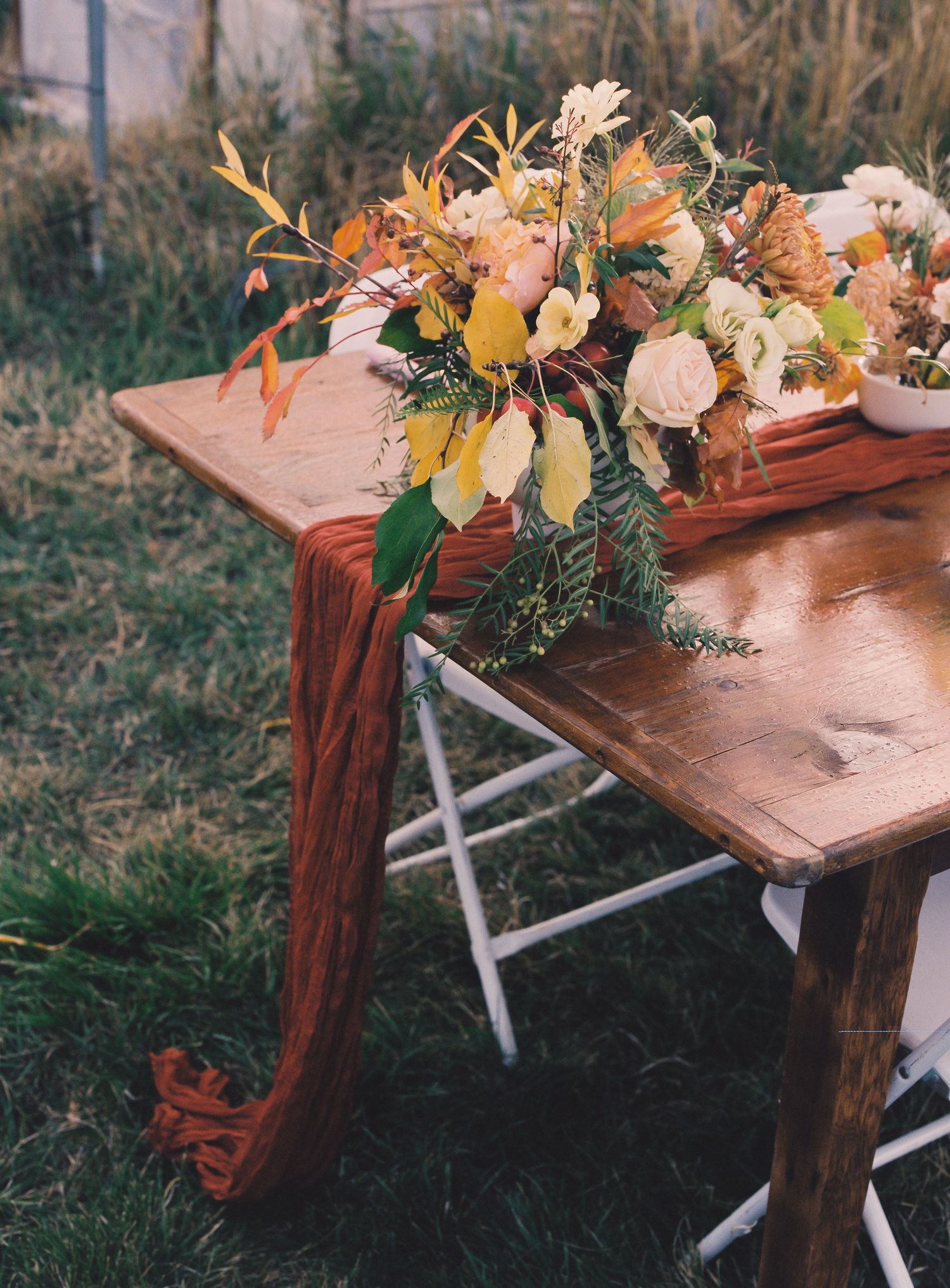 Rustic wooden table with burnt orange fabric runner and autumn floral arrangement outdoors.