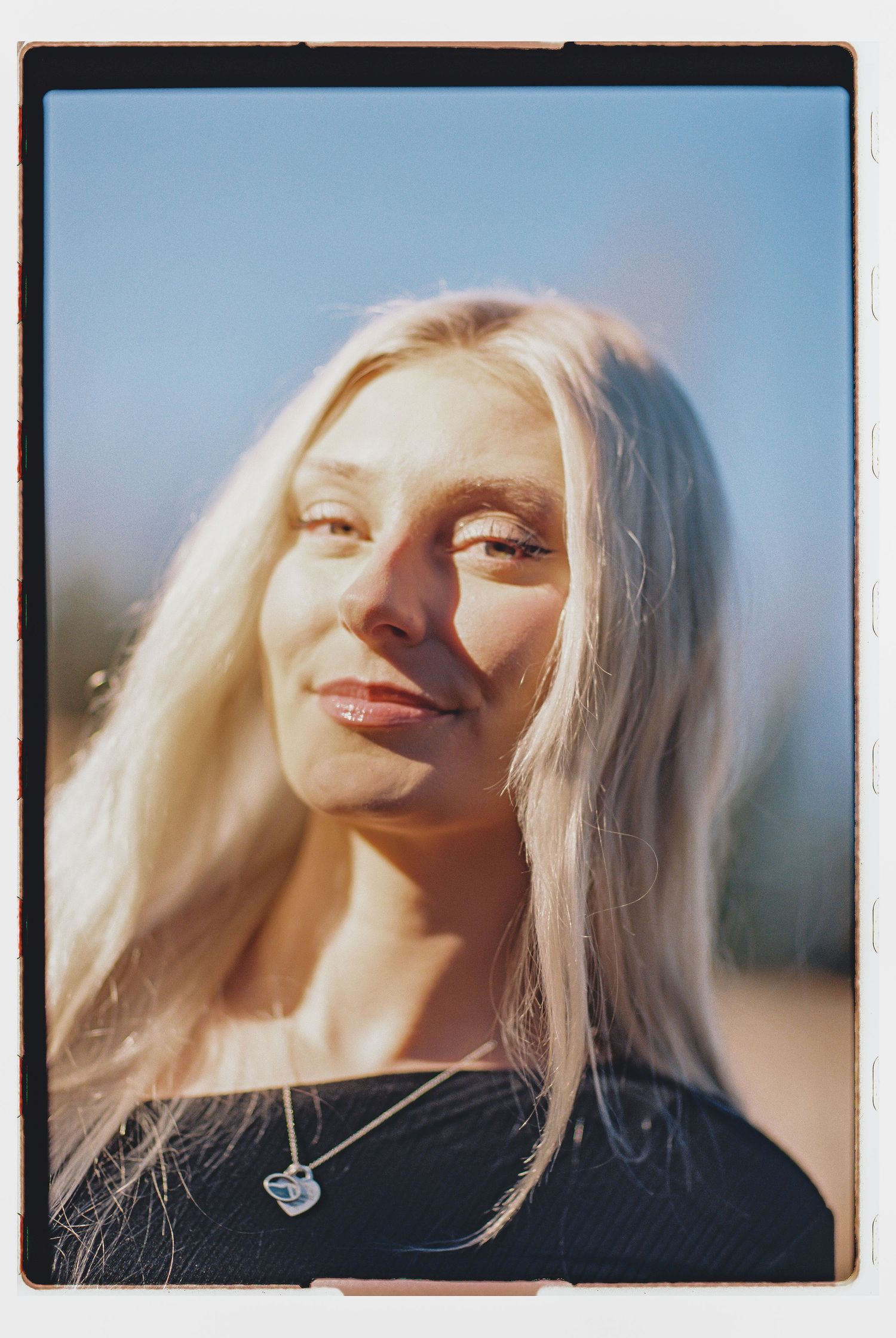 Young blonde woman smiling softly in warm sunlight, wearing a heart necklace and dark top against a blue sky.