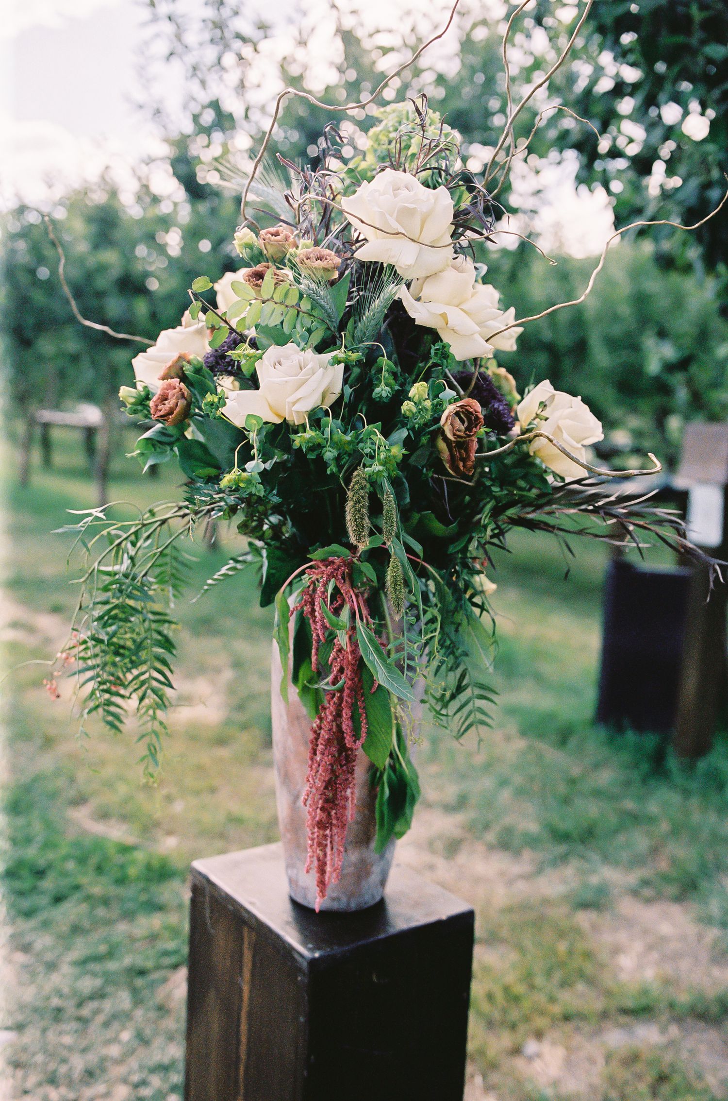 Elegant floral arrangement with white roses, greenery, and amaranthus in a glass vase on a wooden pedestal outdoors.