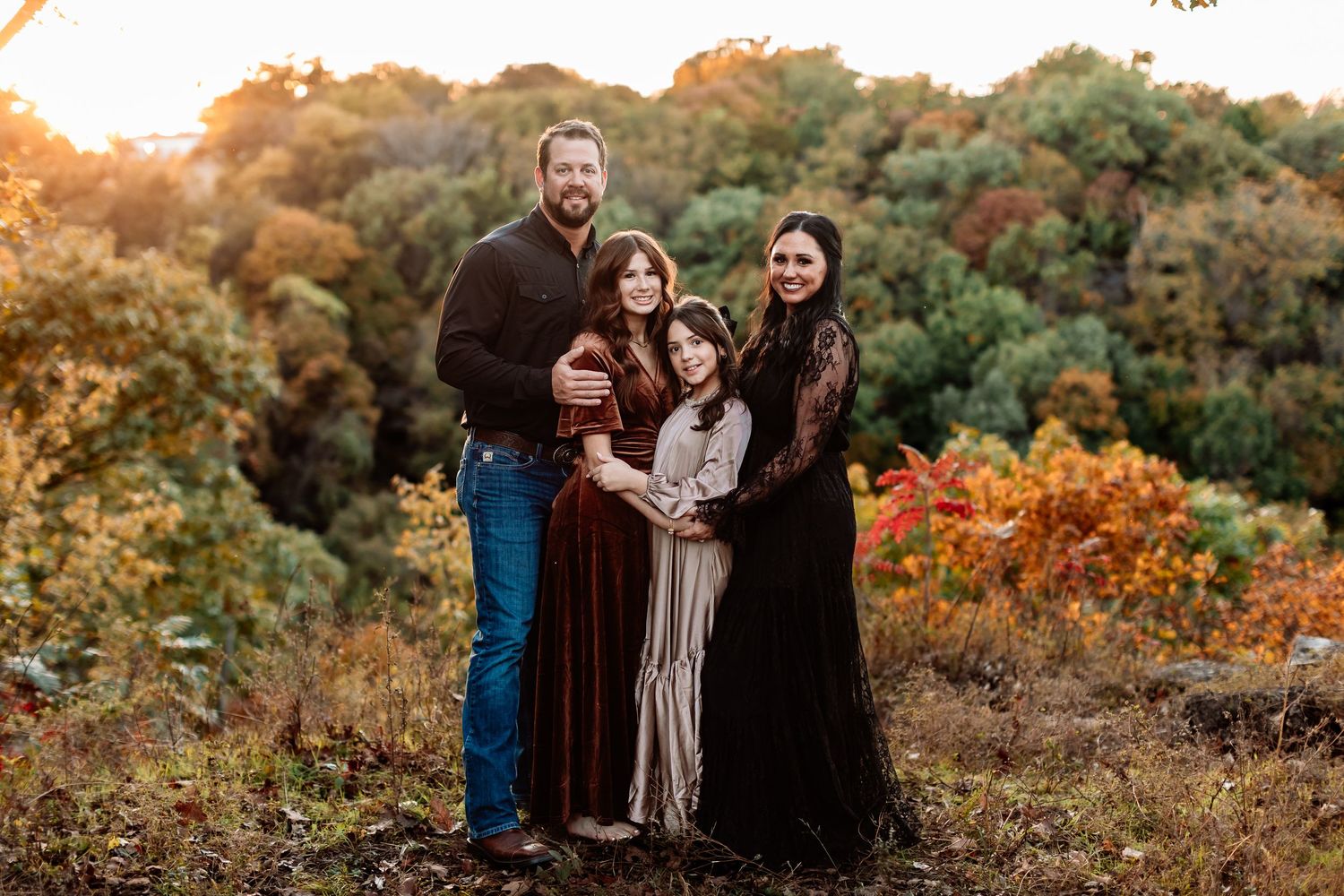 A family stands together on a hillside during golden hour with vibrant autumn foliage in the background.