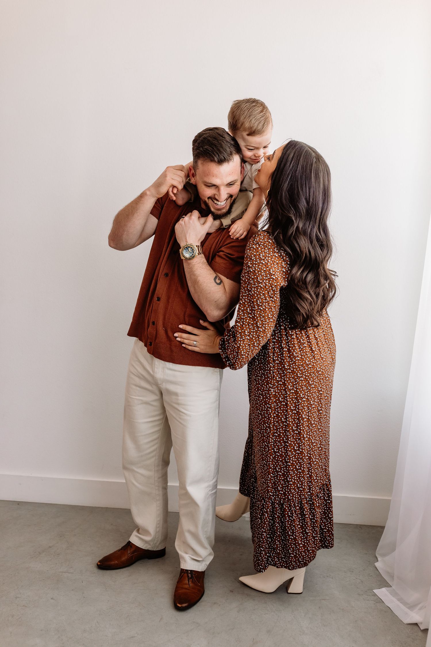 A family shares an intimate moment together against a white wall, with coordinating brown and neutral outfits.