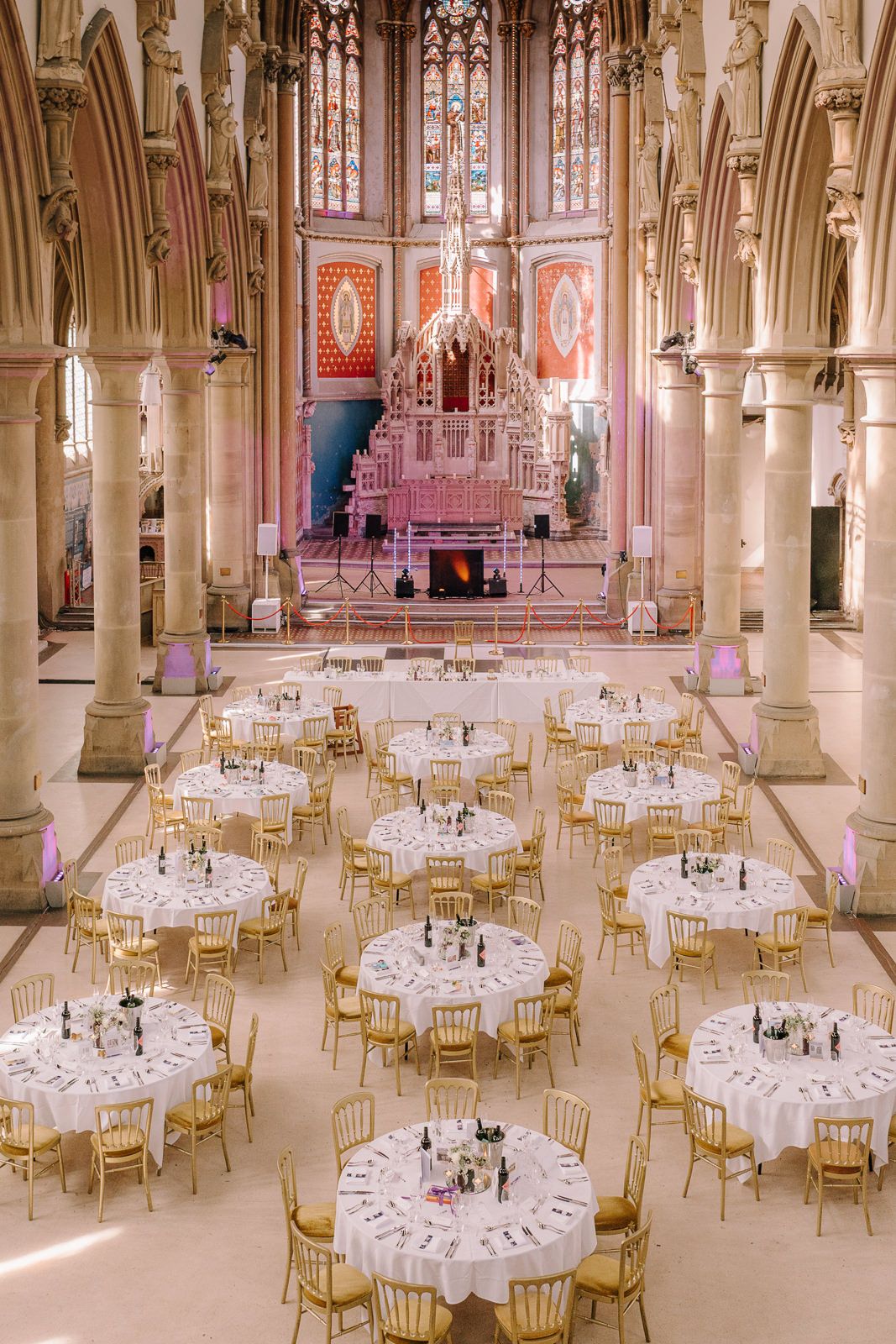 Elegant cathedral wedding reception with round white-draped tables, gold chairs, Gothic arches, and a stunning stained glass altar backdrop.