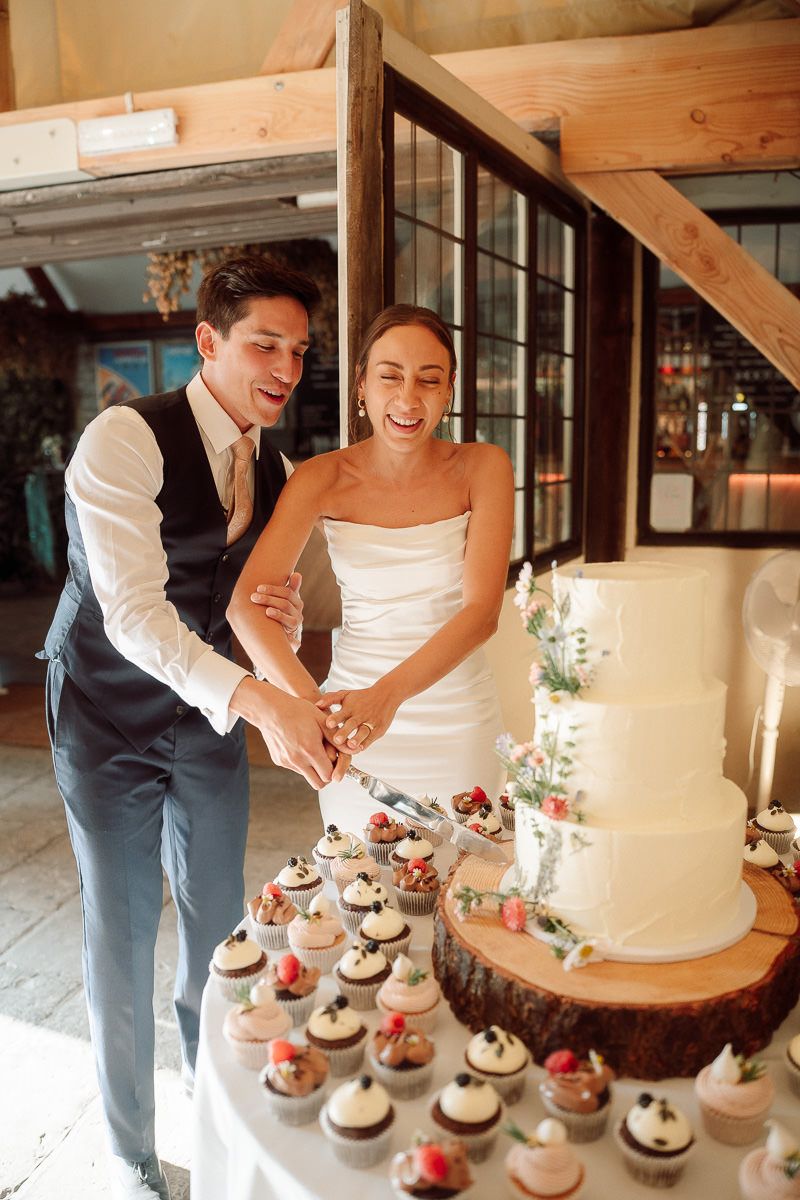 A couple cuts their rustic wedding cake and cupcakes displayed on wooden stands at an indoor reception.