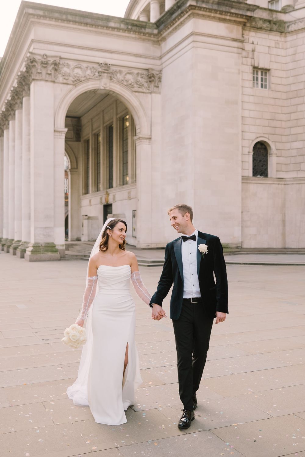 Bride and groom hold hands walking outside a grand neoclassical building, she in a white gown, he in a black tuxedo.