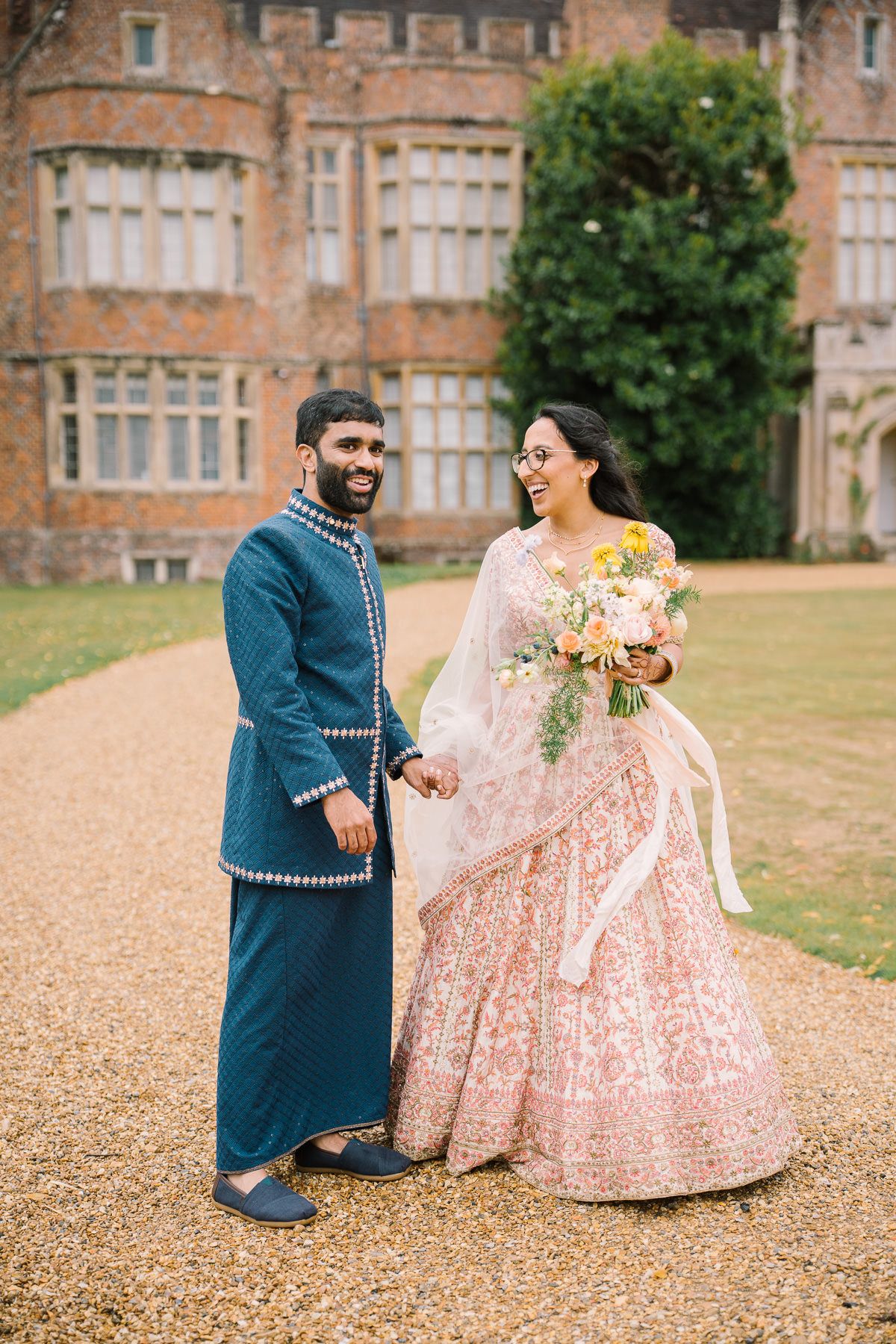 A bride and groom in traditional Indian wedding attire stand together in a garden setting.