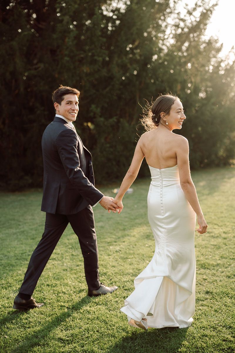 A couple in wedding attire playfully runs and dances together on a sunlit grassy lawn during golden hour.