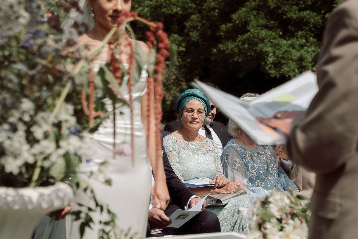 A person in blue sits outdoors at a wedding ceremony with sunlit greenery in the background.