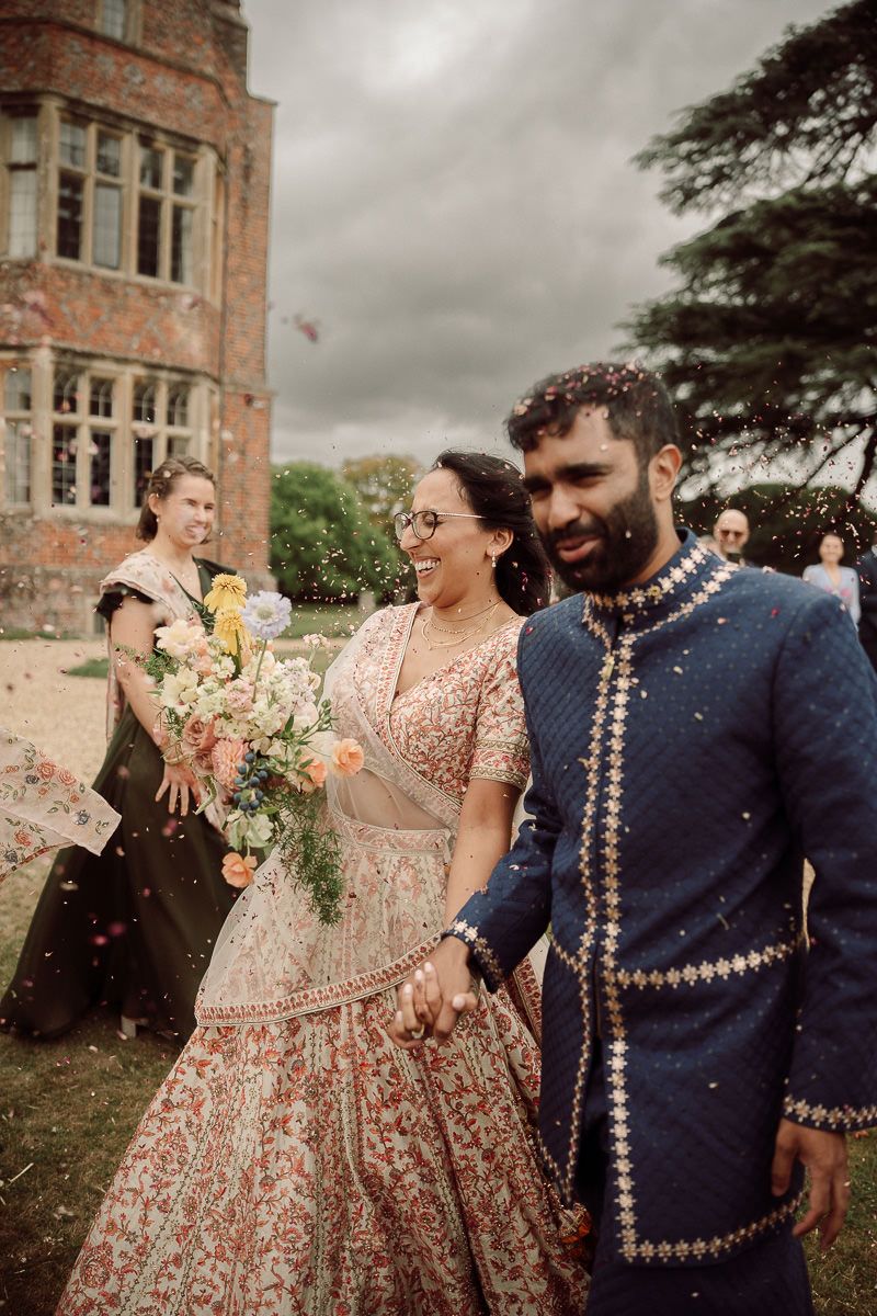 Joyful couple in traditional Indian wedding attire celebrate with guests throwing flower petals at an outdoor venue.