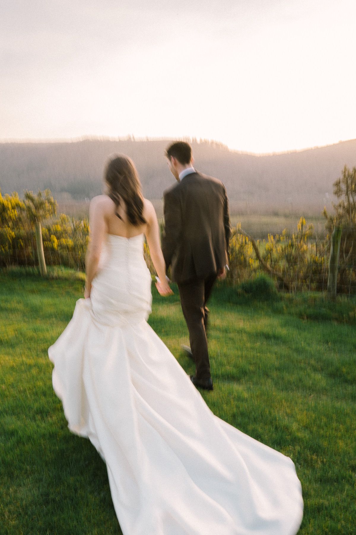 A bride and groom walk together on a grassy field with a scenic mountain view in the background.