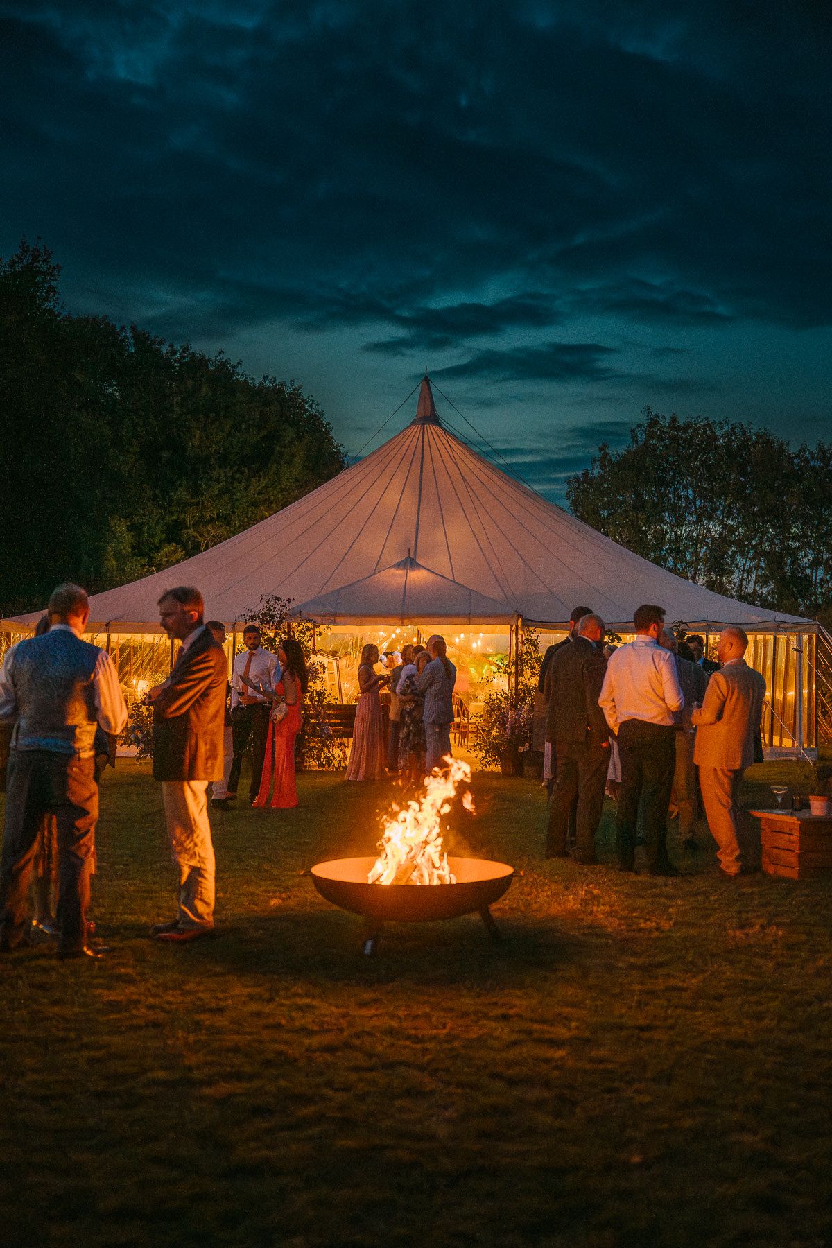 A nighttime outdoor event with a large tent, a fire pit, and people gathered around enjoying the festivities.