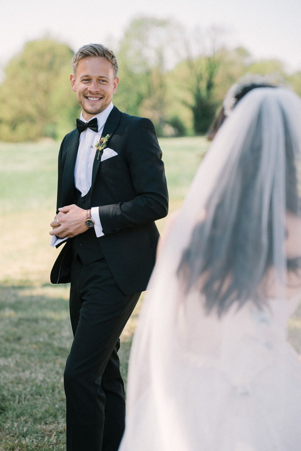 A smiling groom in a black tuxedo stands in a field, facing a bride wearing a long white wedding dress.