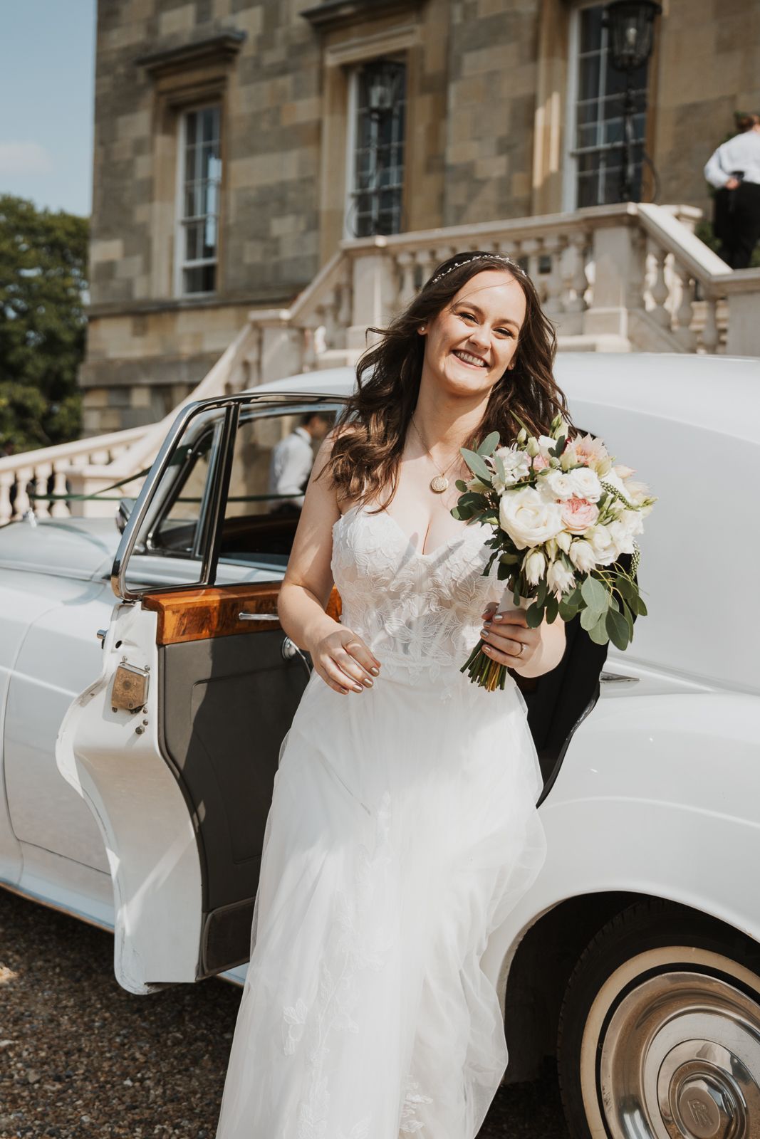 A bride in a white wedding dress holds a pink and white bouquet while stepping out of a vintage white car.