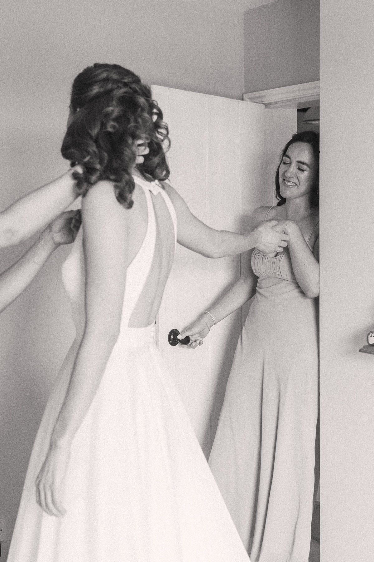 A woman in a white wedding dress stands in a room, with her back to the camera and her curly hair visible.