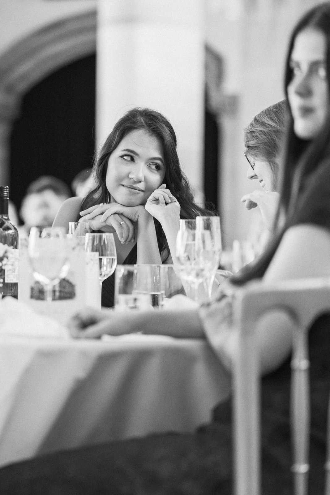 Black and white photo of a woman resting her chin on her hands at a formal dinner table with wine glasses.