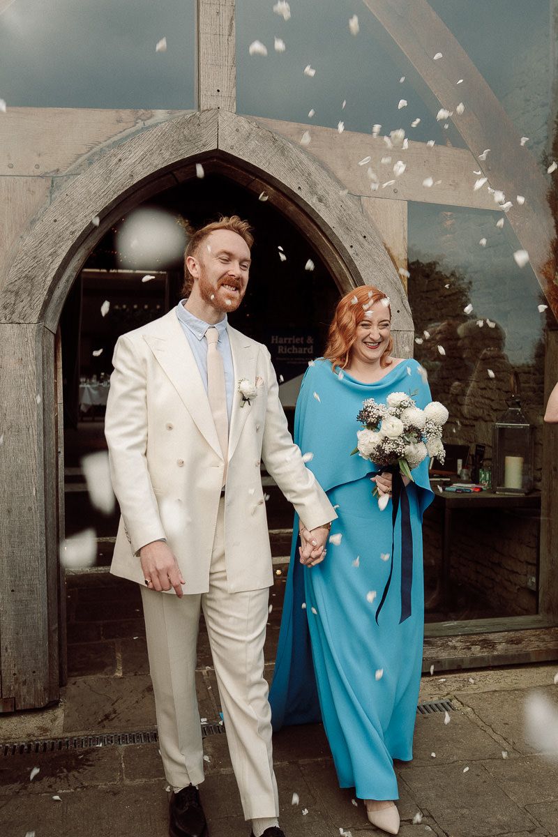 Wedding party exits venue under shower of white flower petals with person in white suit and blue dress walking together.