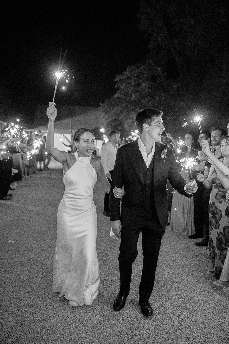 Wedding party exits at night through a sparkler tunnel lined by guests holding lit sparklers creating a magical glow.