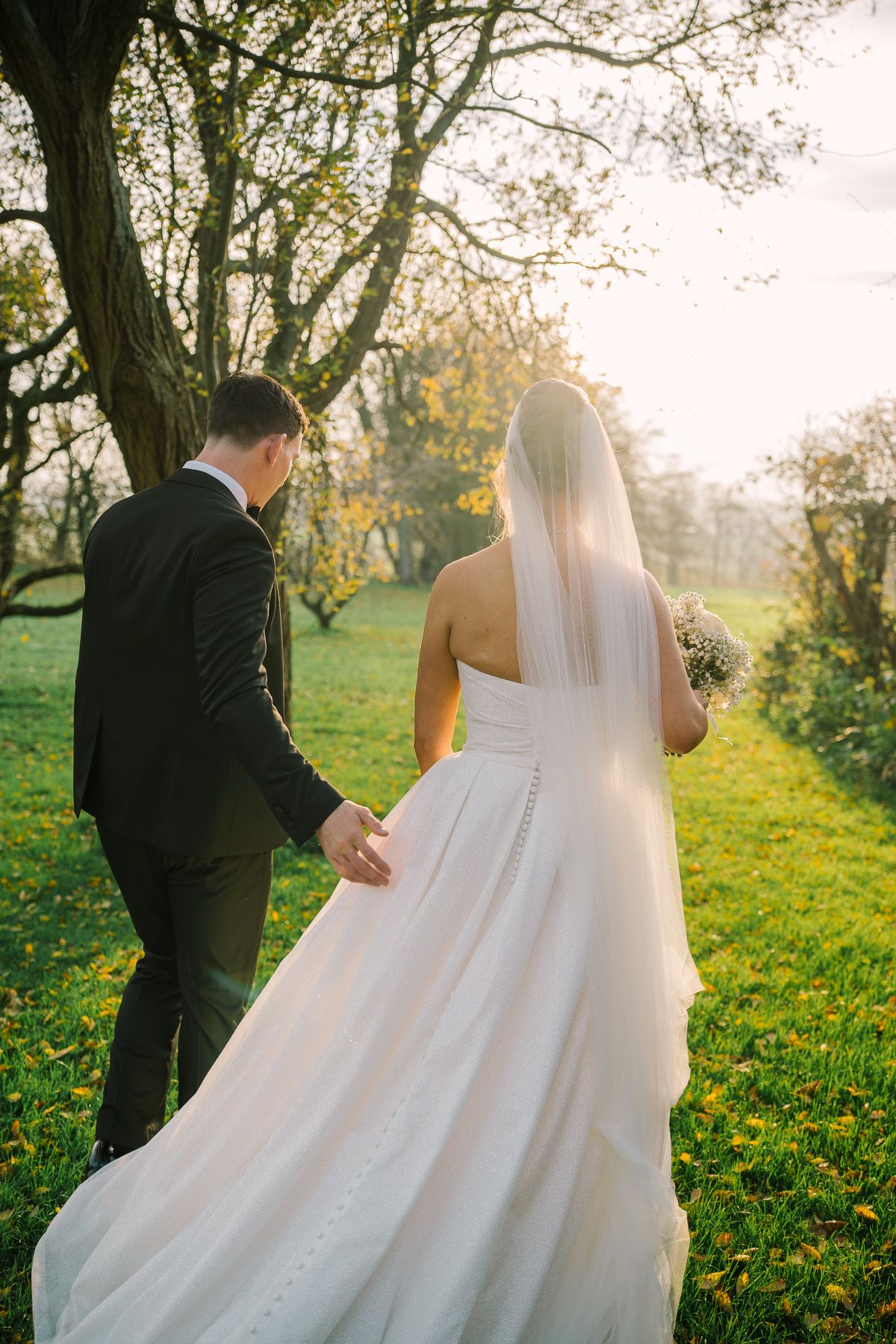 A bride and groom walking in a park on their wedding day, surrounded by trees and greenery.