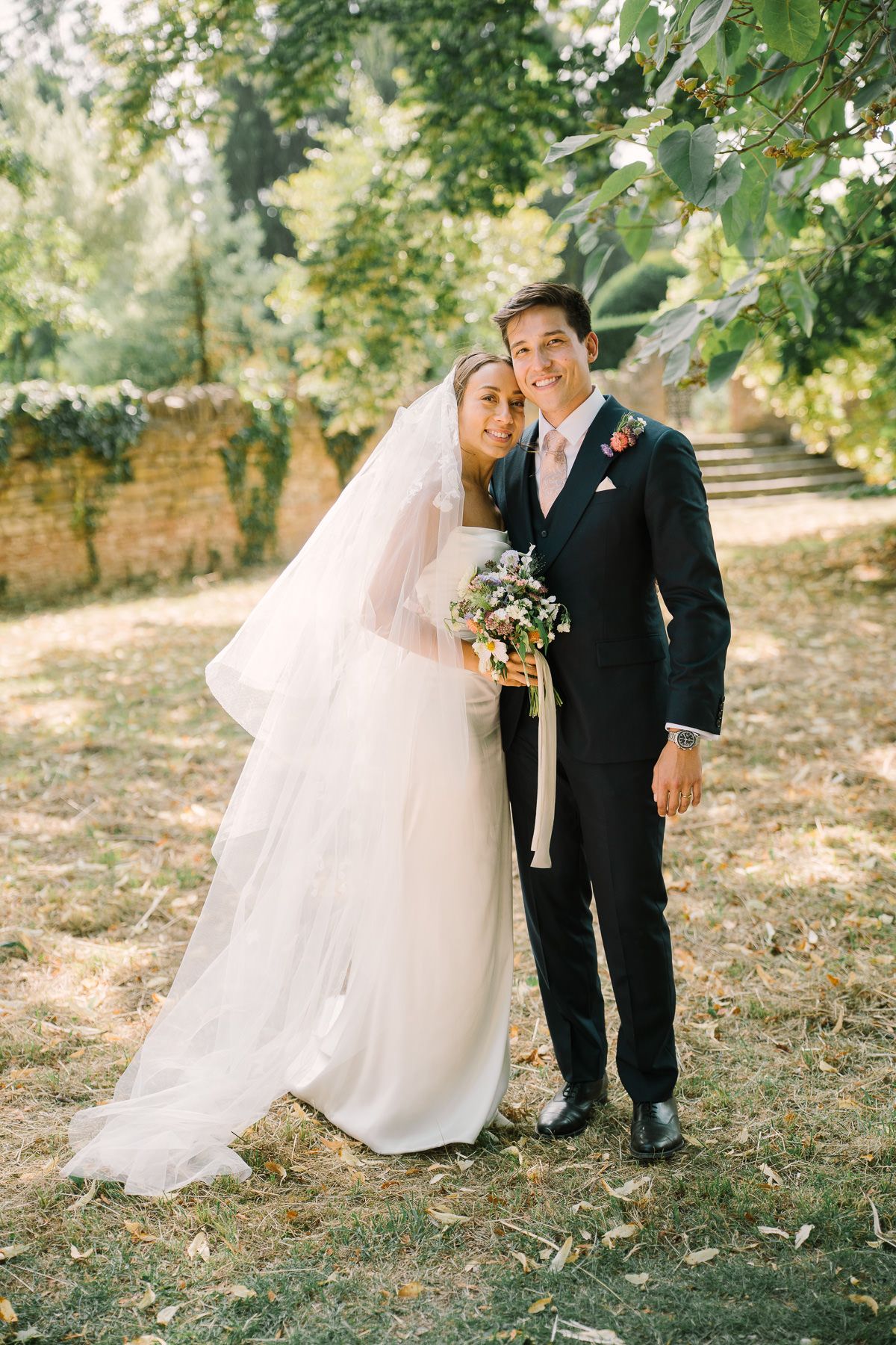 A bride and groom pose together in a garden setting, surrounded by lush greenery and flowers.