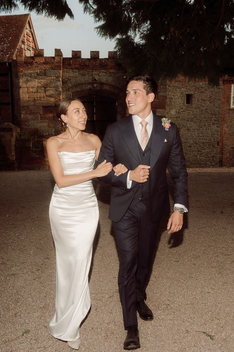 A couple in formal wedding attire walks together along a stone castle wall at dusk.
