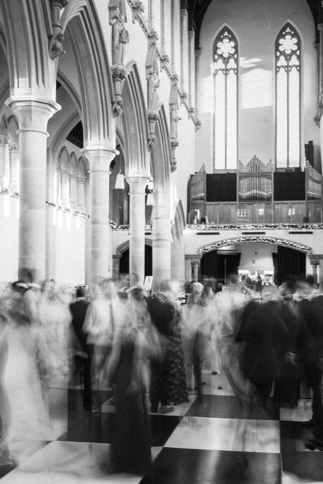Black and white photo of blurred guests moving through a Gothic church interior during a wedding ceremony.