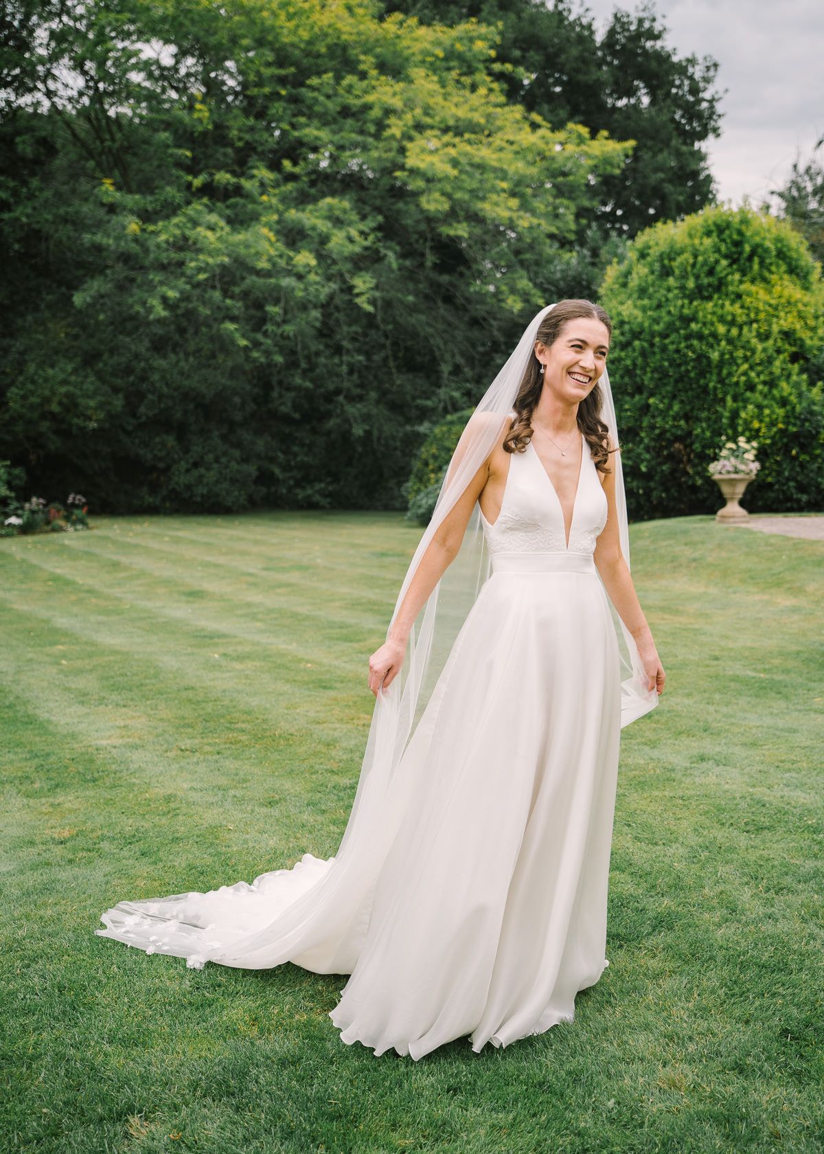A woman in a white wedding dress stands in a grassy field surrounded by trees.
