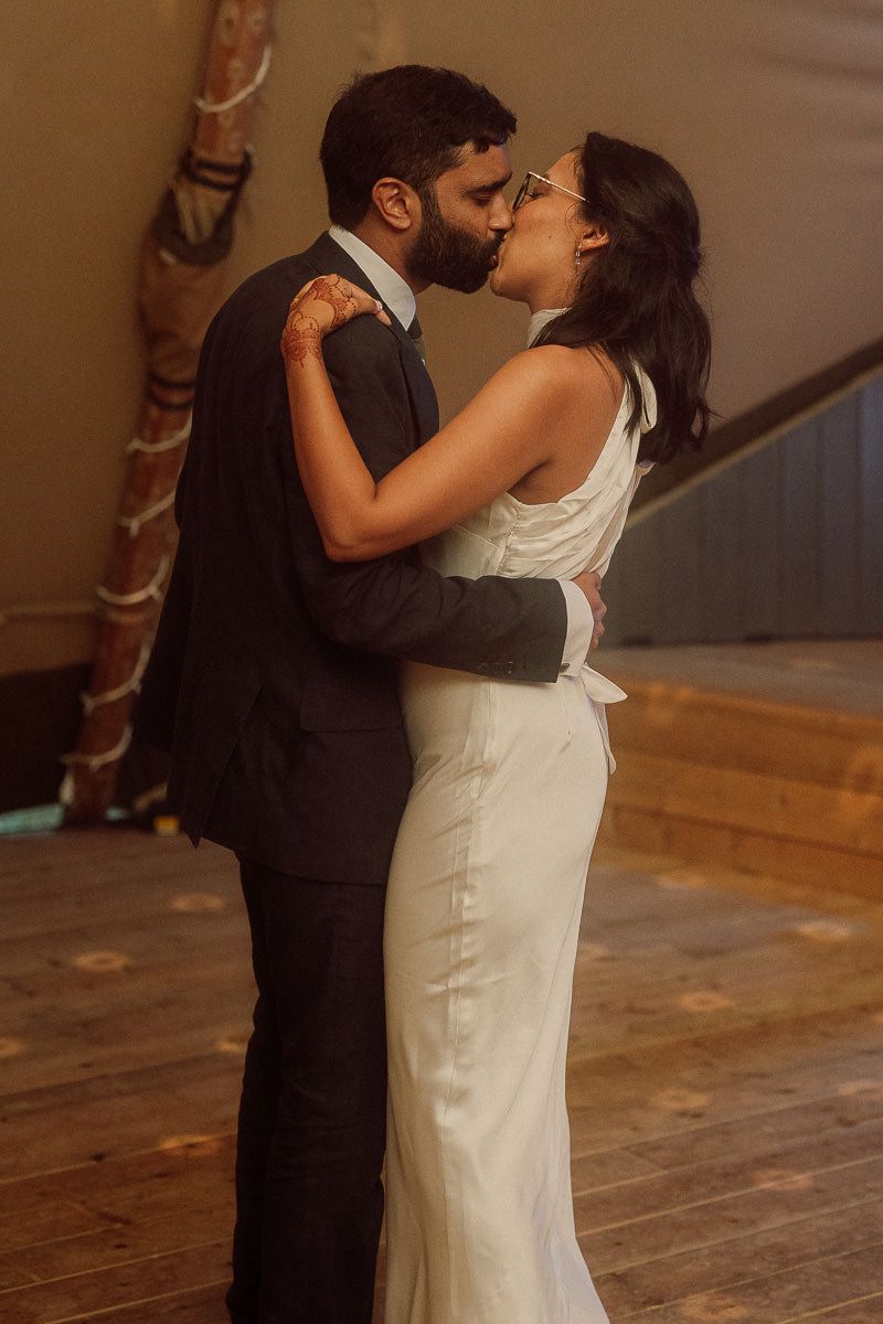 A couple shares a romantic kiss during their first dance at a wedding reception on a wooden dance floor.