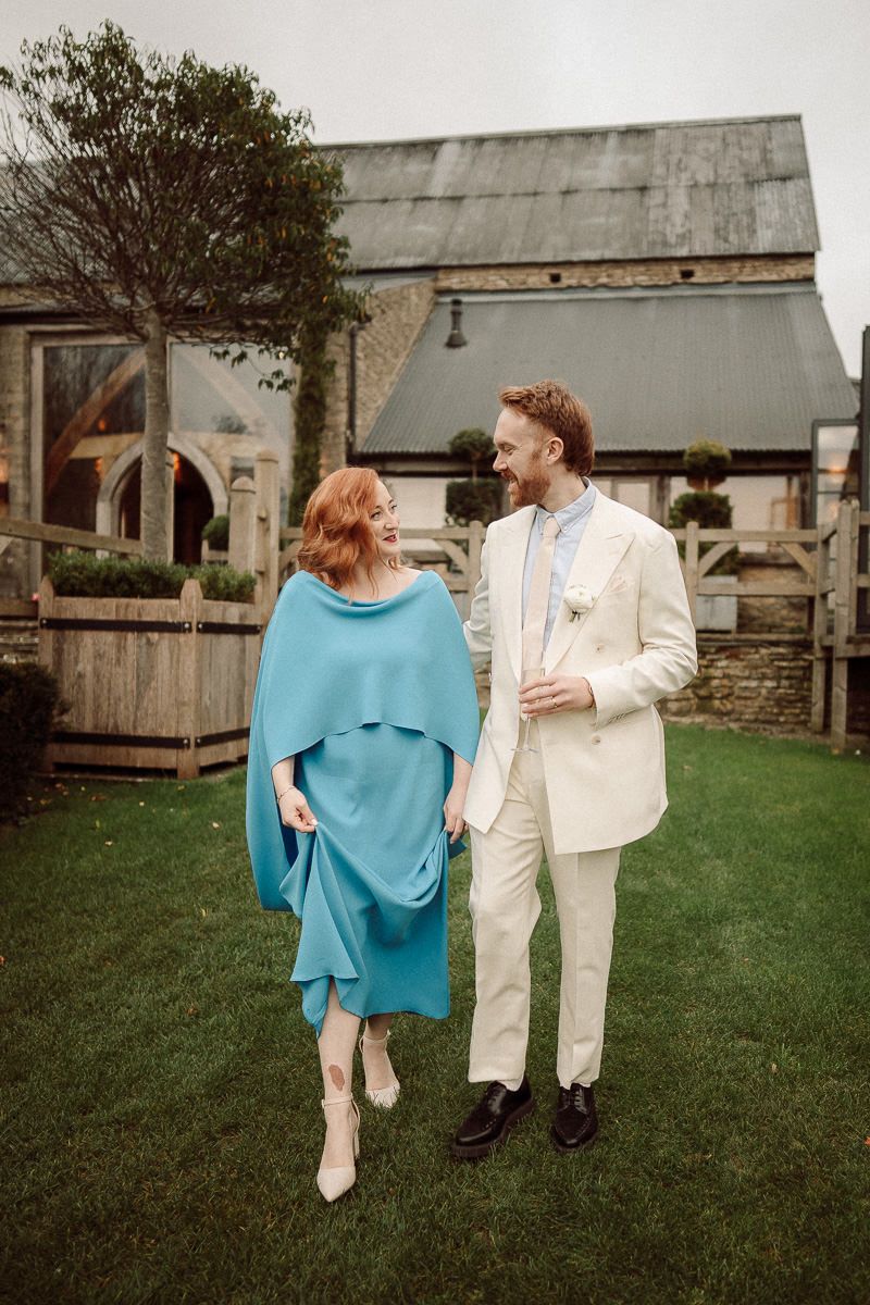 A couple in wedding attire stands together on a grassy lawn near rustic farm buildings.