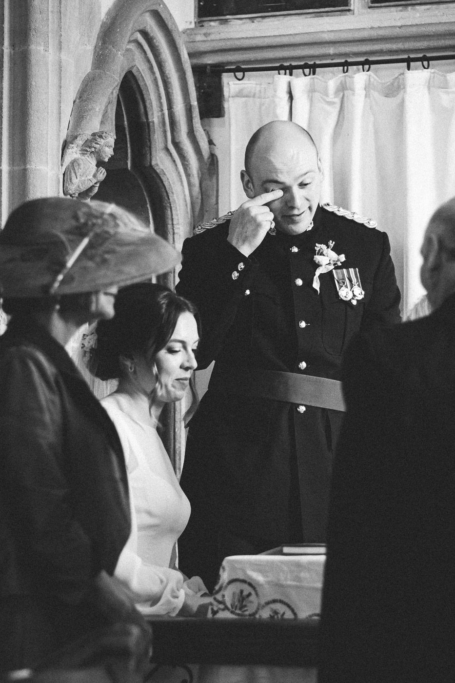 A black and white wedding photo showing a couple in a church with the groom wiping away tears during an emotional ceremony.