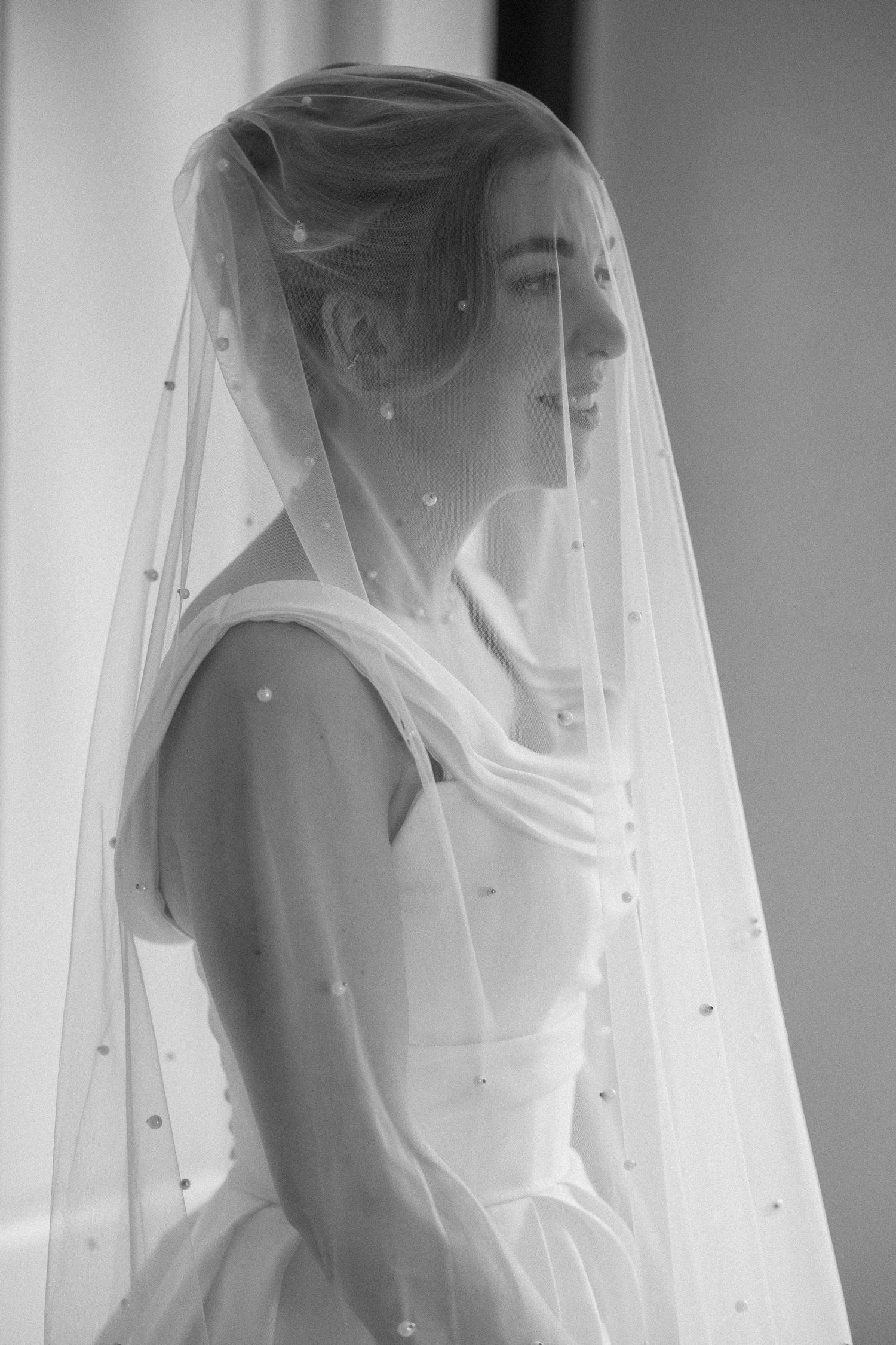 Black and white photo of a smiling bride wearing a pearl-embellished veil and elegant wedding gown.