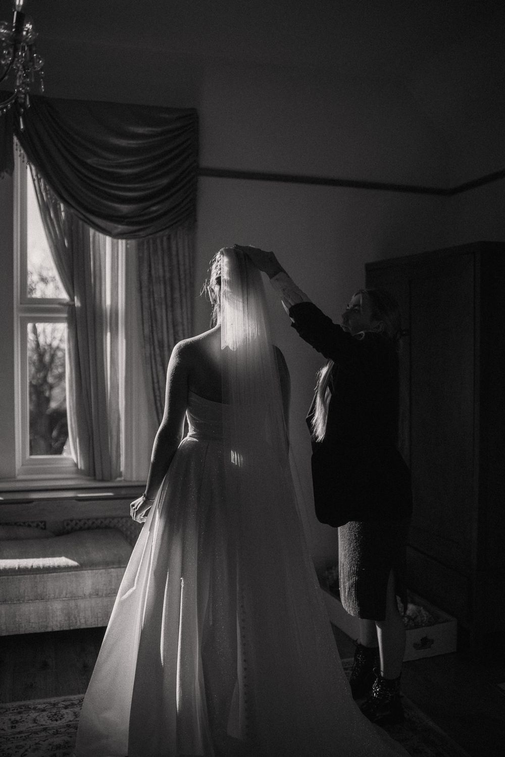 Black and white photo of a bride in a flowing gown having her veil adjusted near a sunlit window.