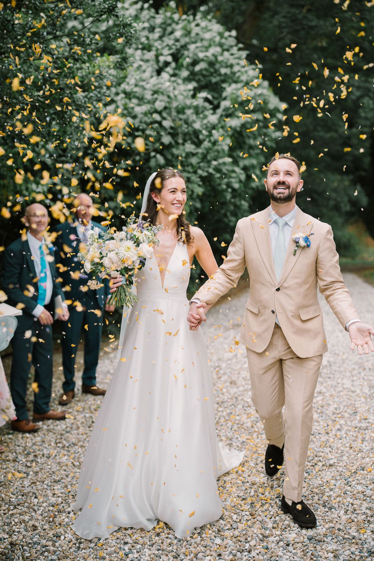 A bride and groom walking together through a flower-filled garden, surrounded by falling petals.