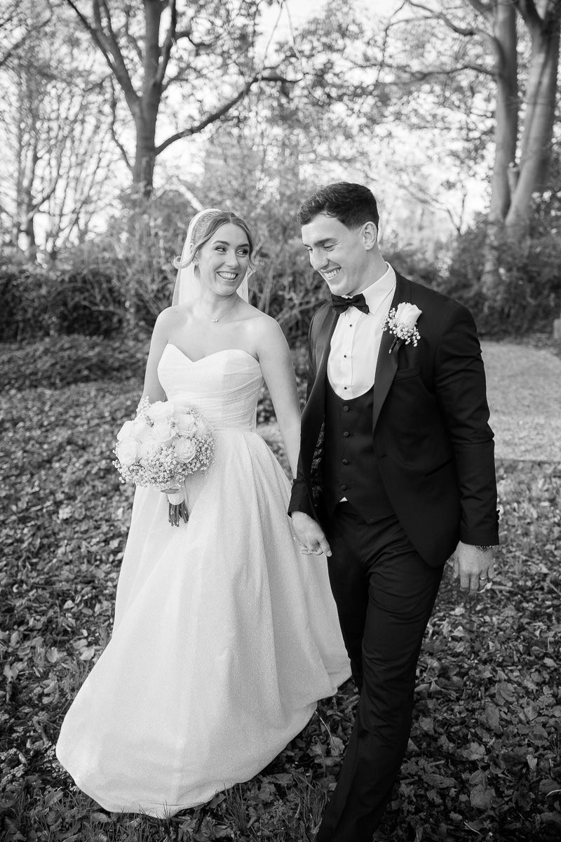 A bride in a white dress and groom in a black suit pose together in a romantic outdoor woodland setting.
