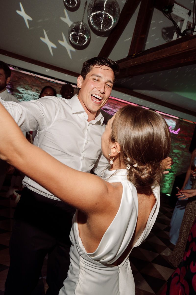 A couple dances and laughs together at a dimly lit party venue with festive lighting in the background.