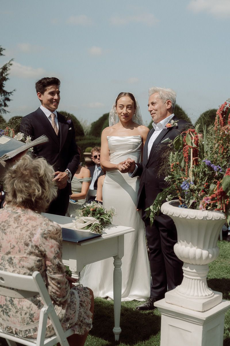 Couple signs marriage documents at outdoor ceremony with decorative white urns and floral arrangements.