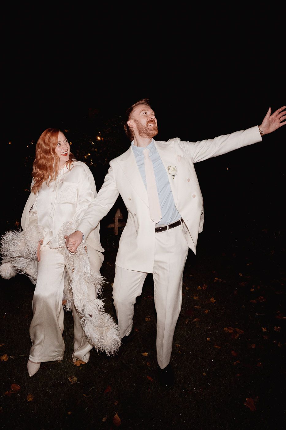 A joyous wedding couple in matching white outfits dance playfully at night with the bride holding feathery details.