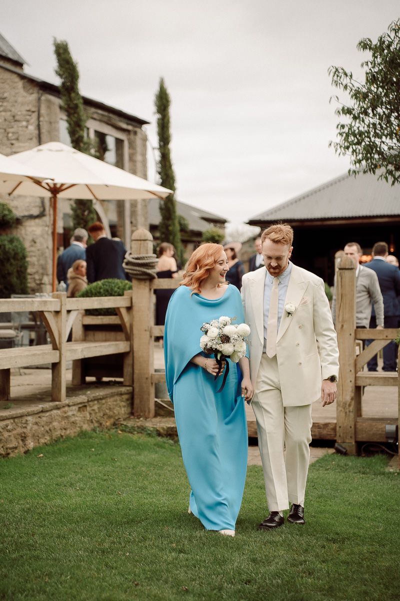 Wedding party members in turquoise and cream attire walk together on a grassy lawn outside a stone building.