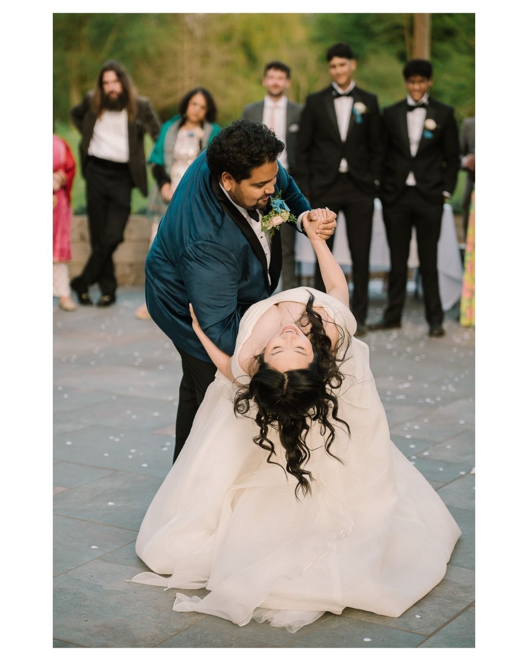 Bride in white gown dips during first dance with groom in teal suit, wedding guests watching outdoors.