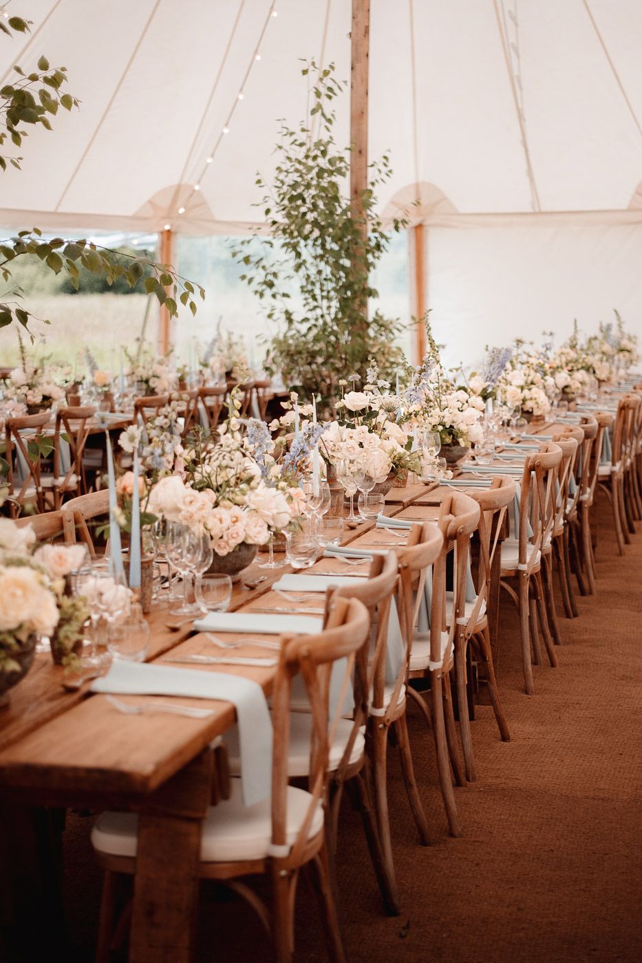 A wedding reception tent interior with elegant long wooden tables set with glassware, linens and floral centerpieces.
