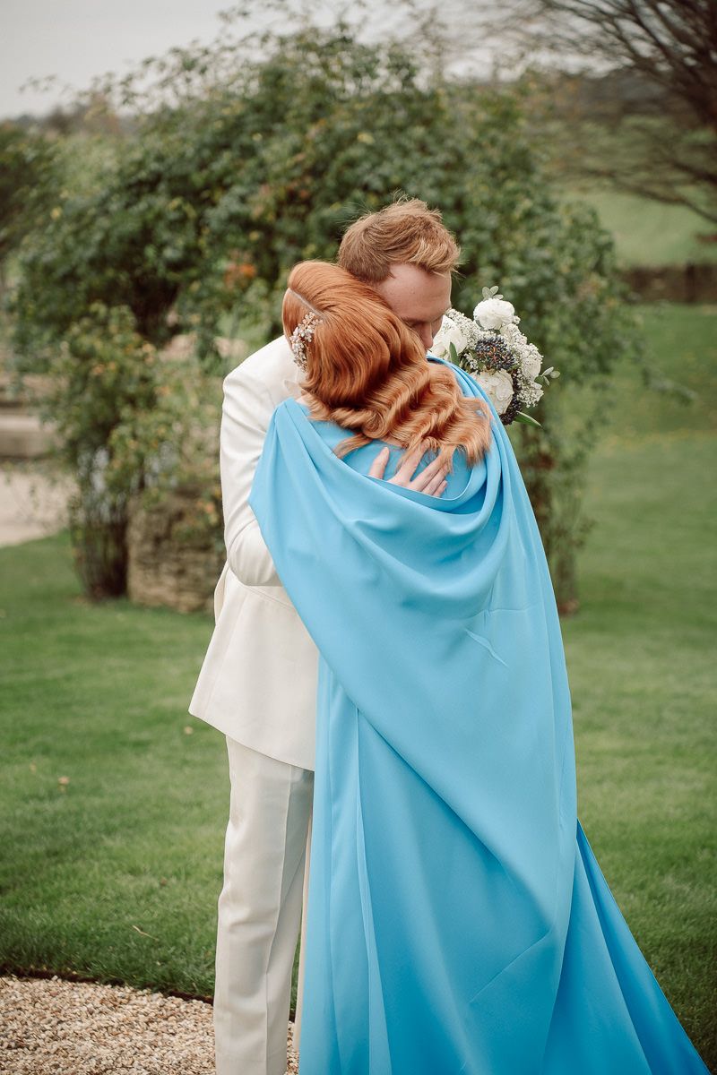 A couple shares an intimate moment wrapped in a flowing light blue cape against a green outdoor backdrop.