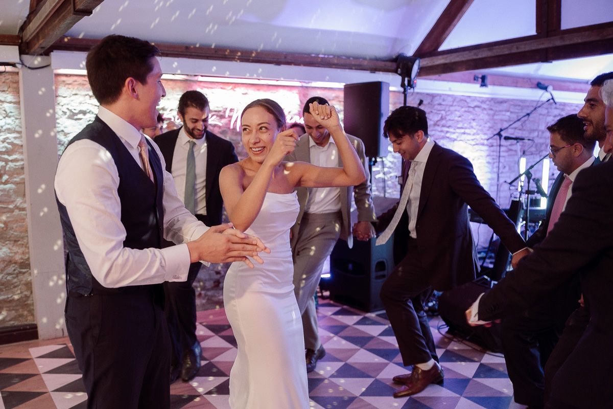 Wedding guests dance and celebrate together on a checkered floor at an indoor reception venue.