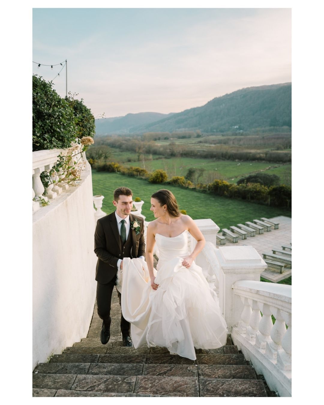Bride and groom walk up white stone steps at elegant outdoor wedding venue with scenic mountain valley views.