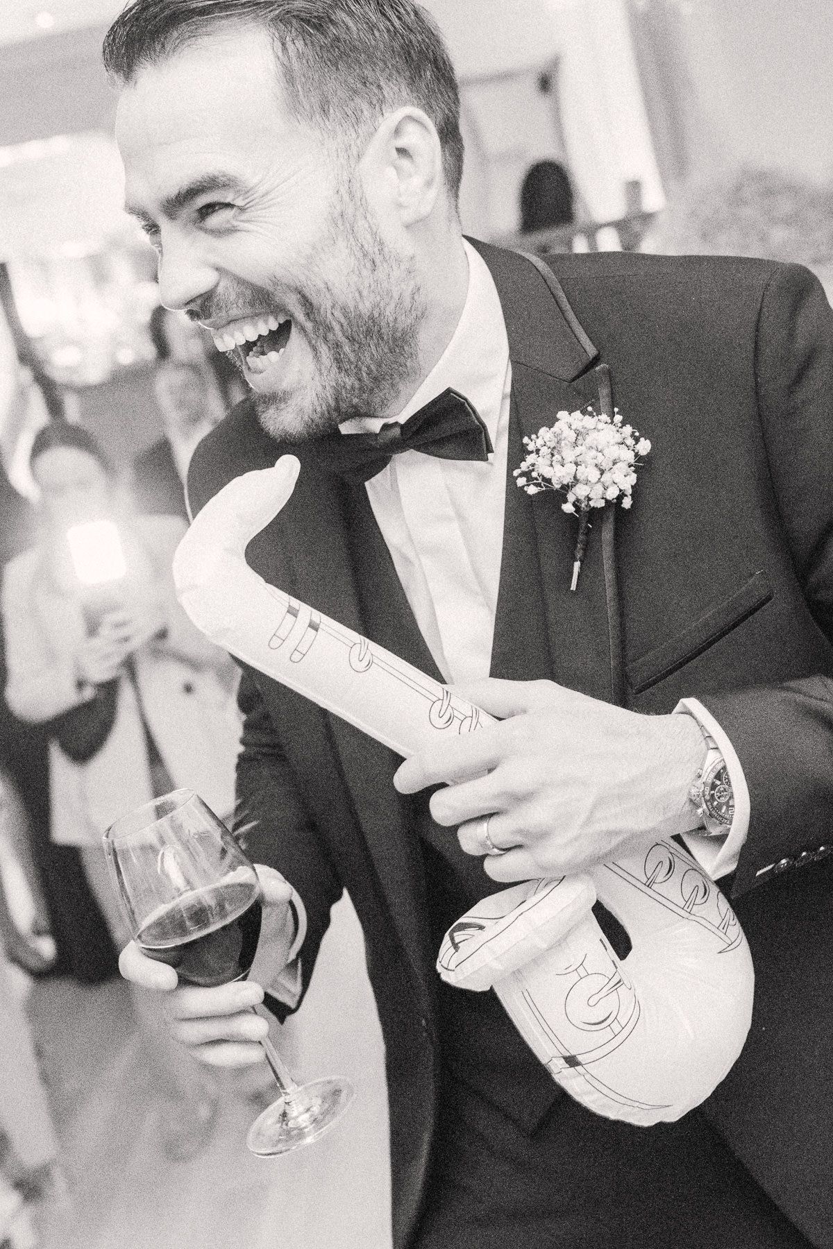 A smiling man in a tuxedo holds a guitar and glass of wine, likely at a wedding or formal event.