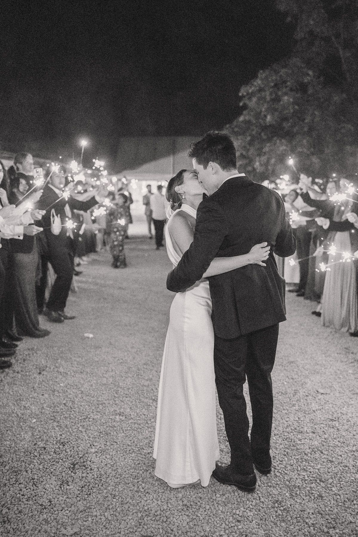 A black and white image of a bride and groom embracing at their wedding, surrounded by cheering guests holding sparklers.