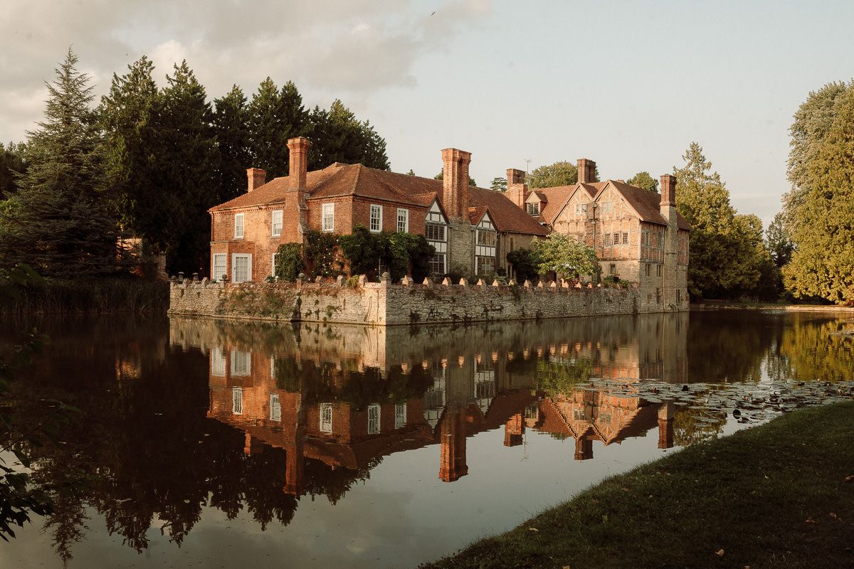 Historic brick mansion with multiple chimneys reflects perfectly in still lake waters at sunset.