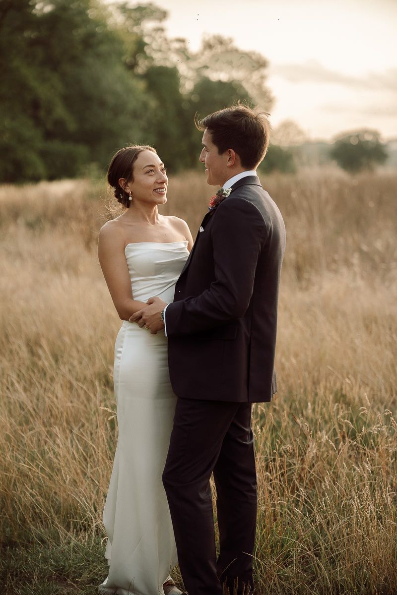 A couple in wedding attire embraces in a golden field at sunset with trees in the background.