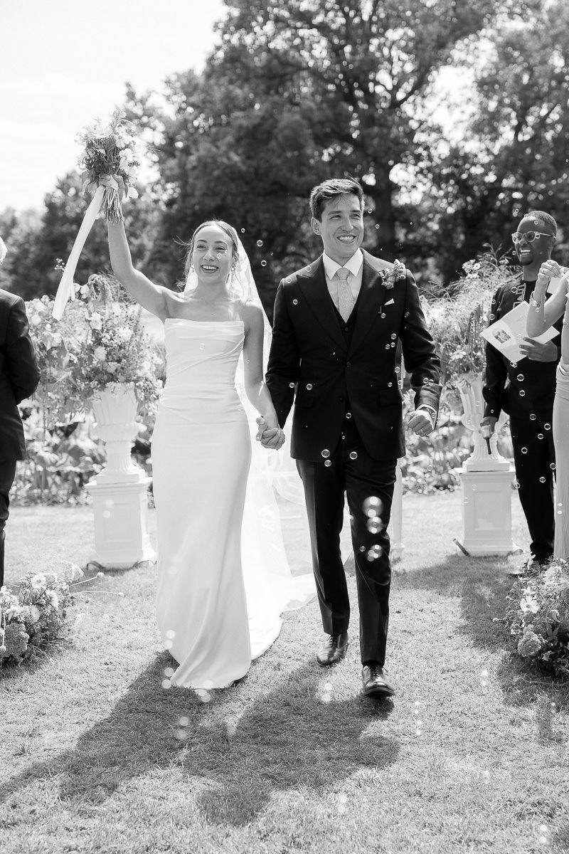 A bride and groom walk down an outdoor garden path while guests shower them with flower petals in black and white.
