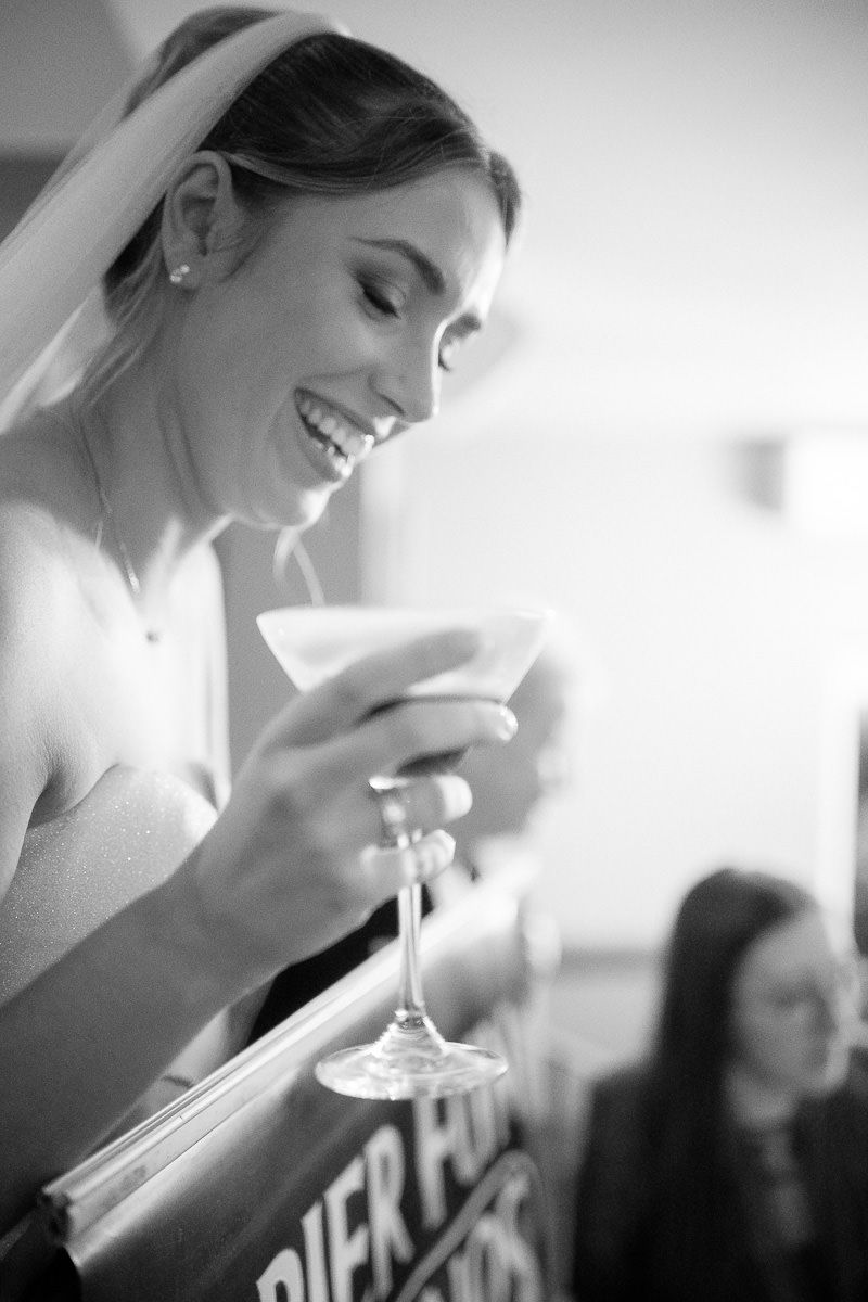 A bride in a white dress smiles while holding a martini glass during a wedding celebration.