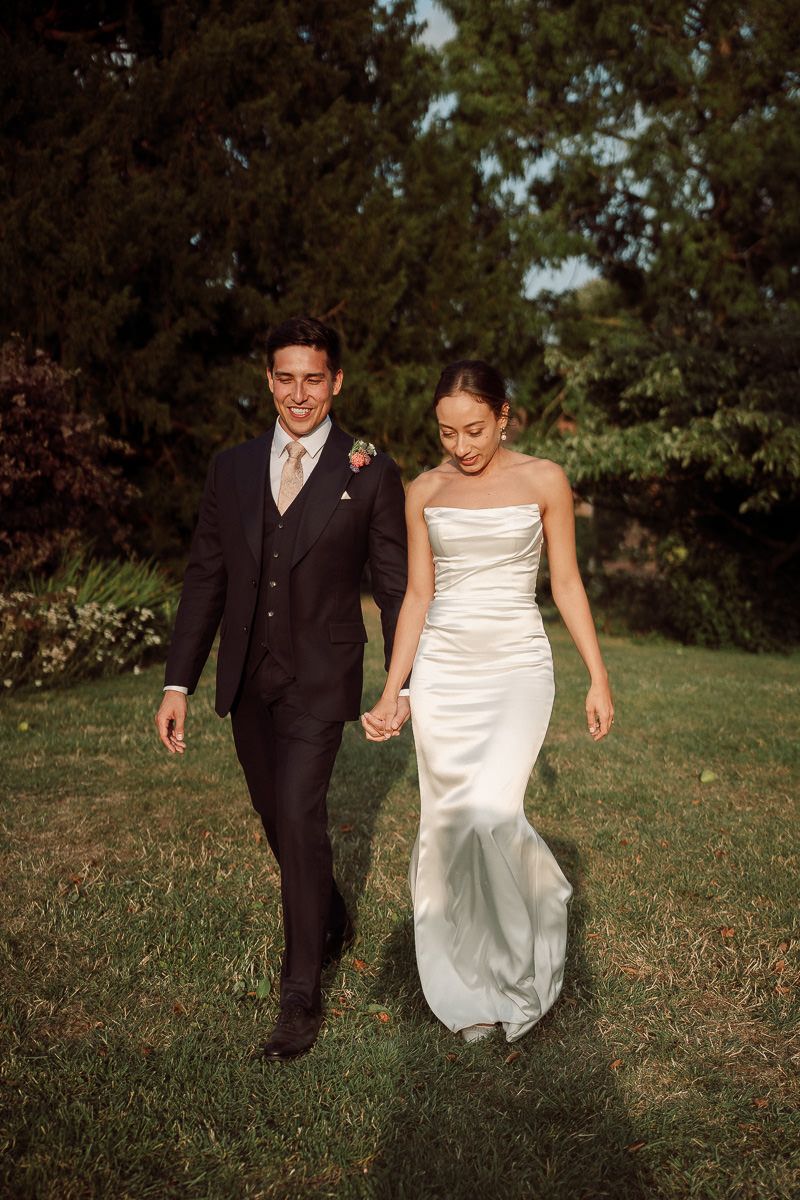 A couple walks hand in hand on their wedding day, with the bride in a white satin gown and groom in a dark suit.